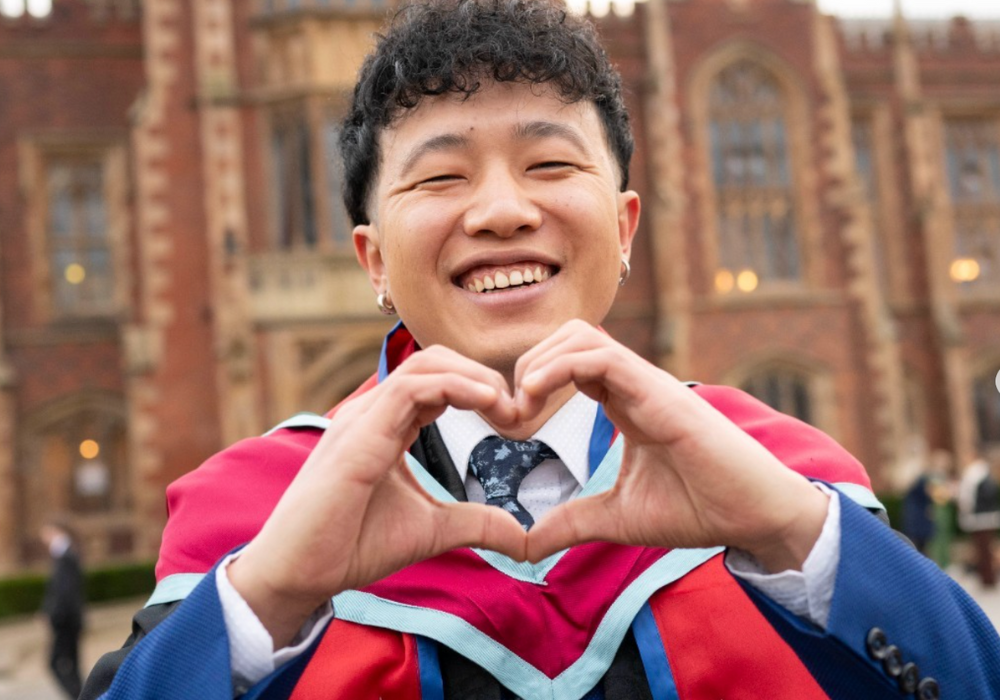 A young man in a graduation gown forming a heart shape with his hands.